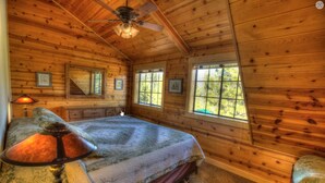 Une chambre à coucher dans une cabane en bois, avec un lit, une lampe, un ventilateur de plafond et des fenêtres donnant sur la forêt.