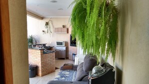 A living room with a grey sofa, a coffee table, and a hanging fern. A kitchen with a microwave and a brick stove is visible in the background. - Edicula Foz (Foz do Iguaçu)