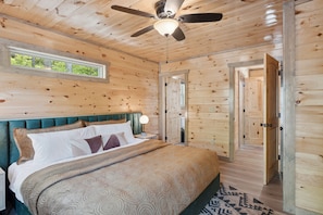 Un dormitorio de cabaña de madera con cama, ventilador de techo, ventana y puerta que da a otra habitación.