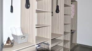 Un armario de madera con múltiples estantes y espacio para colgar, una cesta con artículos de aseo y un perchero metálico con una tetera.