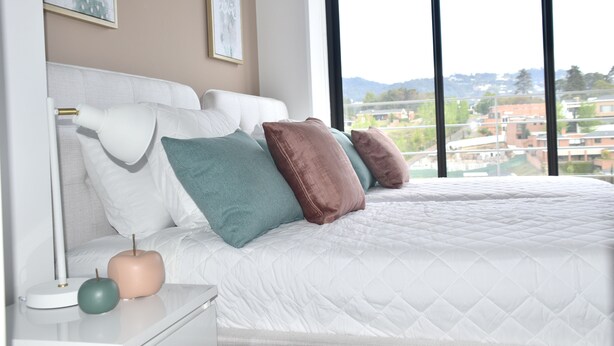 Un dormitorio con una cama grande, ropa de cama blanca y vistas a un paisaje urbano a través de una ventana.