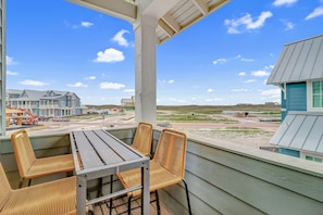 A balcony with wicker chairs and a table, overlooking a building with a blue roof and a view of a road with vehicles. - Dazzling home with a balcony and updated appliances - pool, gym, & on-site golf (Port Aransas)