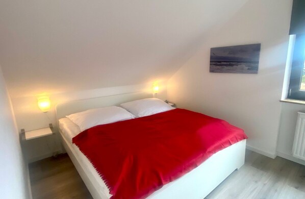 Una cama individual con ropa blanca y una manta roja en una habitación con paredes blancas y una ventana.