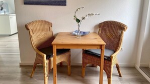 Een eethoek met twee rieten stoelen en een houten tafel, een vaas met bloemen, en een schilderij van een zeegezicht aan de muur.