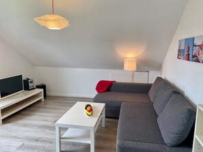 Ett modernt vardagsrum med en grå soffa, ett vitt soffbord, en platt-TV och en lampa.