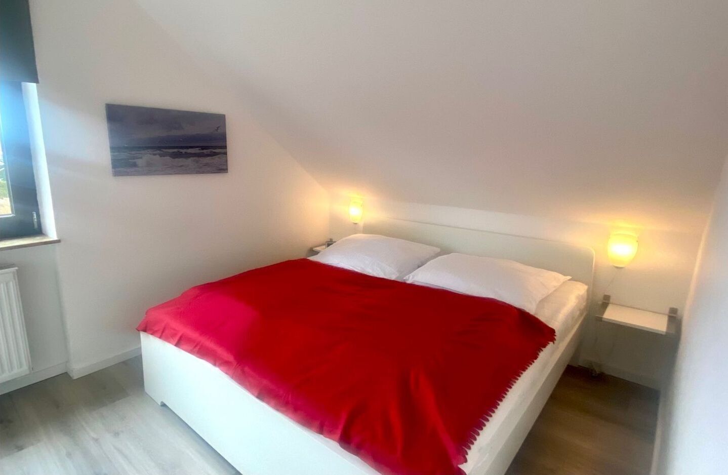 Ein Schlafzimmer mit einem Bett, einer roten Decke, weißen Kissen, einem Nachttisch mit Lampe und einem Bild an der Wand.