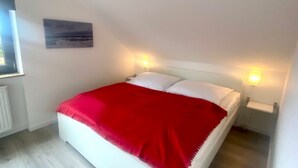 Een slaapkamer met een bed, een rode deken, witte kussens, een nachtkastje met een lamp en een schilderij aan de muur.