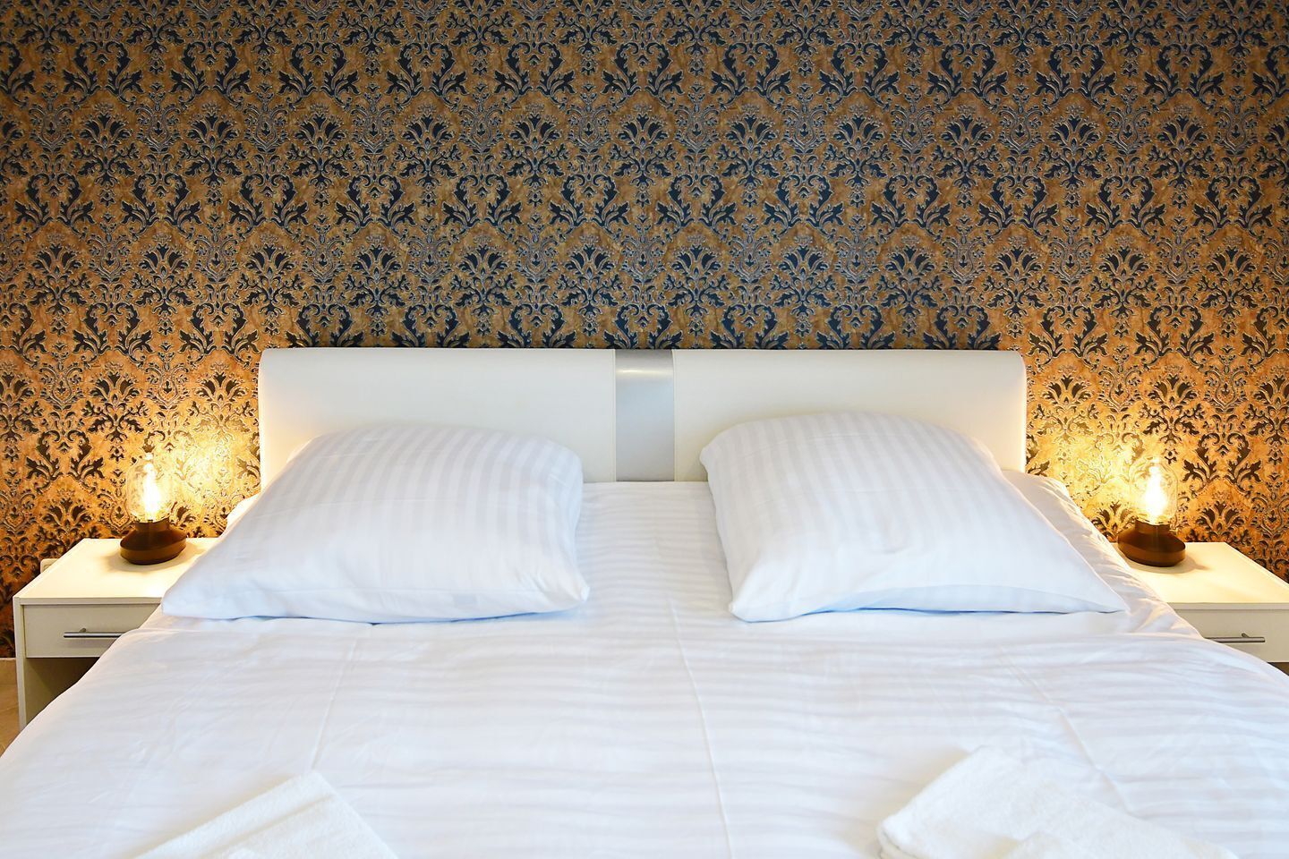 Una cama doble con sábanas blancas, dos mesitas de noche con lámparas y un papel tapiz estampado.