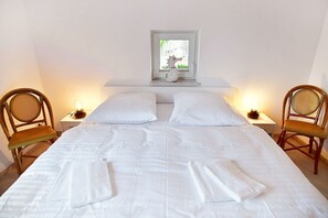 Un lit double avec du linge de lit blanc, deux fauteuils en osier, une table de chevet avec une lampe et une fenêtre donnant sur un paysage verdoyant.