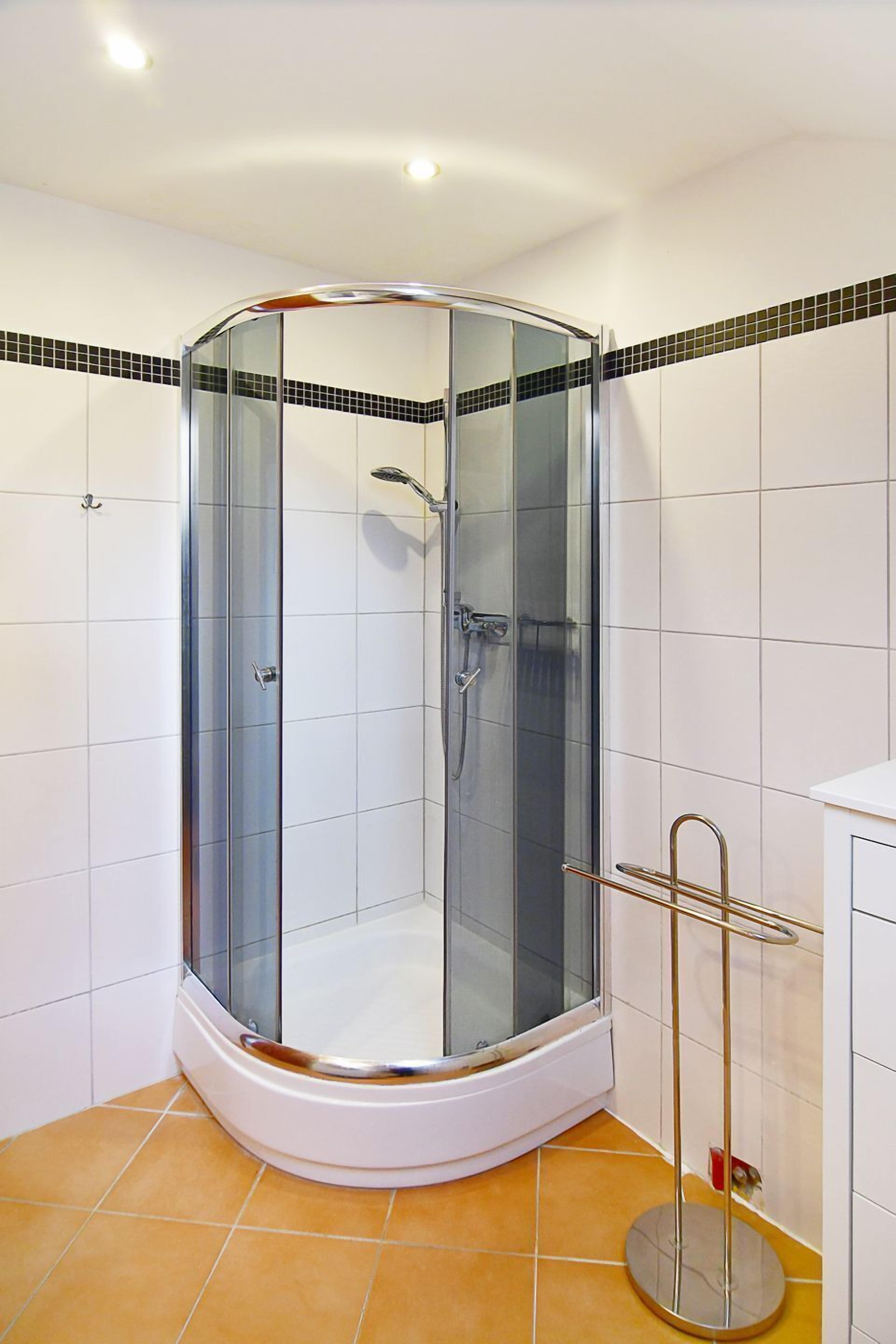 Ducha en esquina con mampara de vidrio, paredes de azulejos blancos y lavamanos en esquina con grifo dorado.