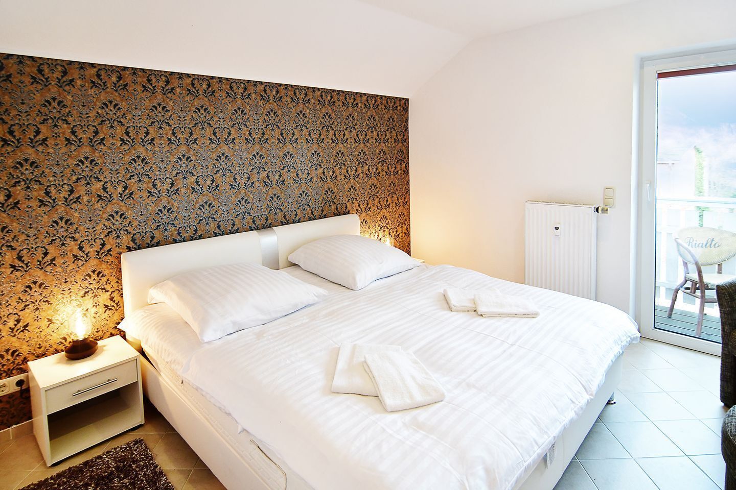 A neatly made bed with white linens and pillows, a bedside table with a lamp, and a patterned wallpaper on the wall.