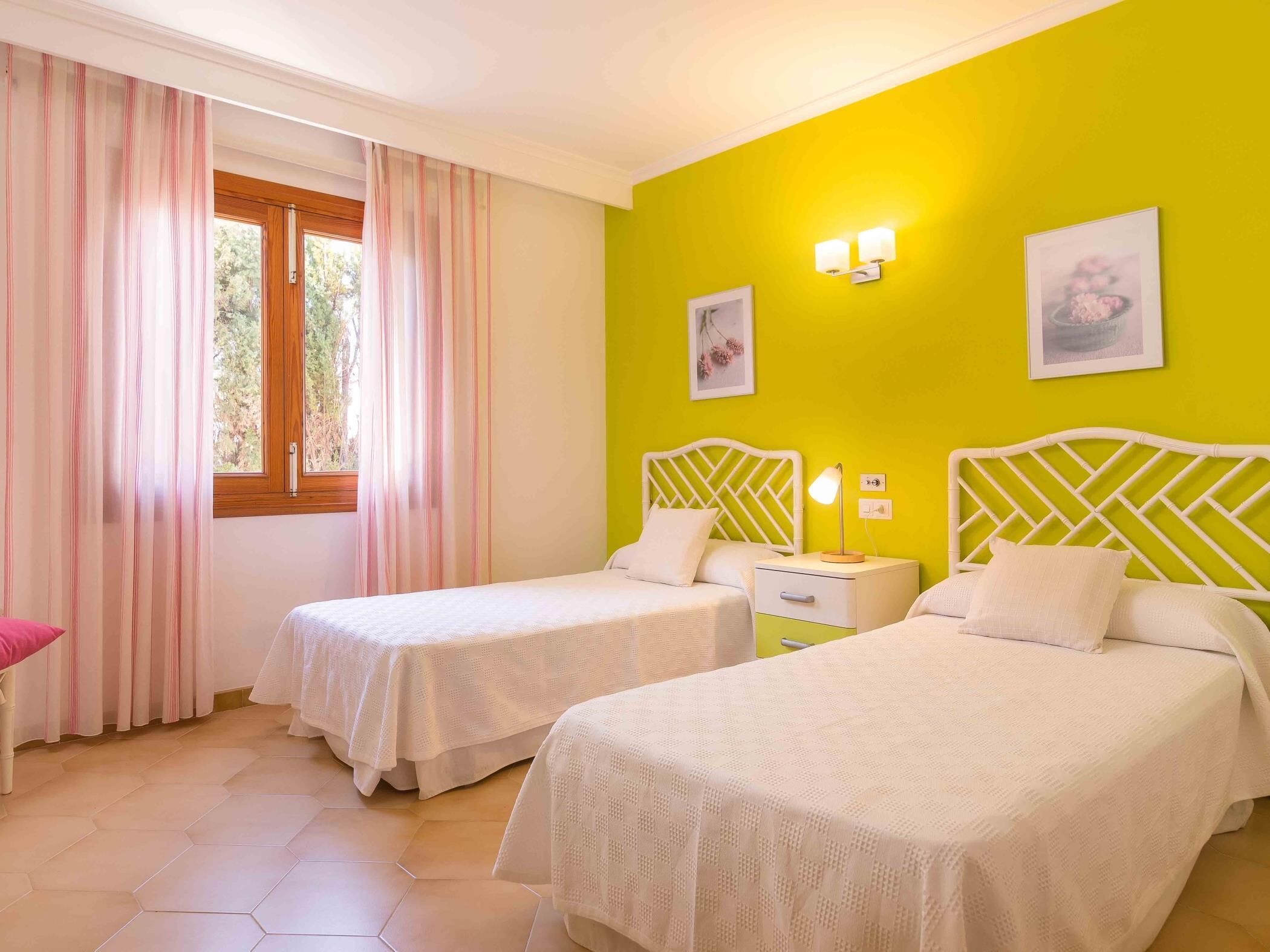Une chambre avec deux lits, une fenêtre avec des rideaux, une table de chevet avec une lampe et deux lampes fixées au mur.