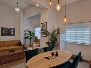 A modern living room with a round wooden table, green upholstered chairs, a yellow sofa, and a large window with blinds.