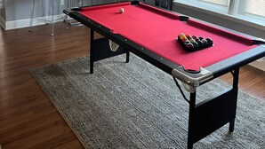 Une table de billard dans une pièce dotée d’un tapis gris, d’une fenêtre avec des stores et d’une chaise avec un coussin.