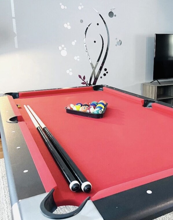 A pool table with a red felt surface, a set of pool cues, and a rack of billiard balls.