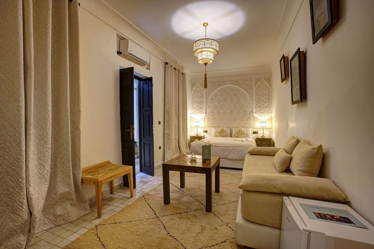 A hotel room with a bed, a wooden bench, a small table, a chandelier, and a wall with framed pictures.