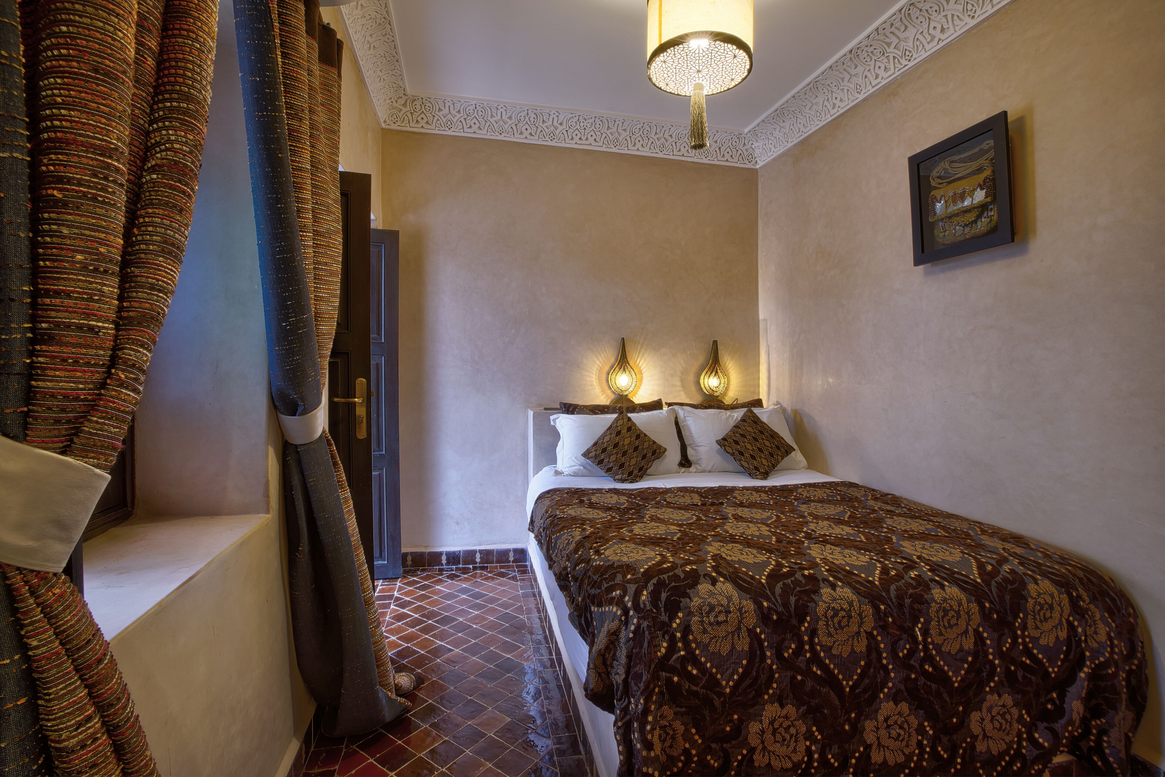 A bedroom with a patterned bedspread, two wall-mounted lamps, and a framed picture on the wall.