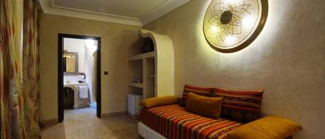 Habitación de hotel con sofá, cojines estampados, una lámpara de pared decorativa, alfombra estampada y vista al baño.