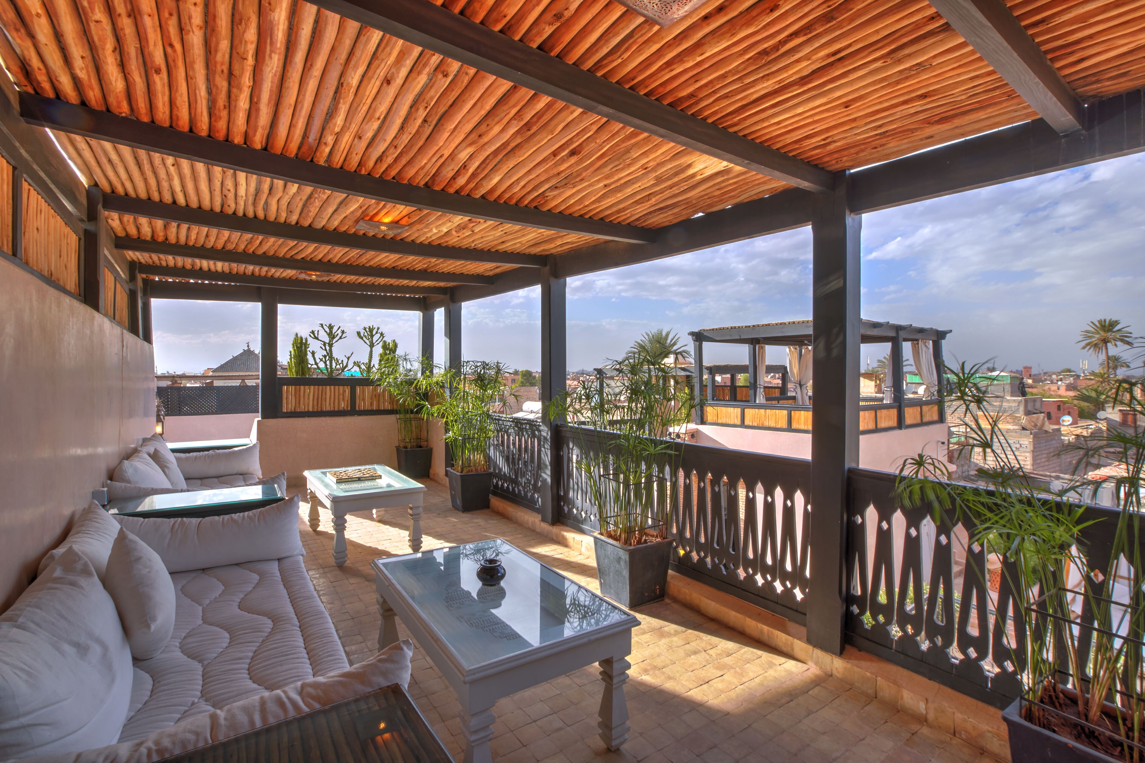 A covered outdoor seating area with a sofa and a table, surrounded by potted plants and overlooking a cityscape.