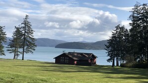 A rustic cabin with a sloped roof, situated on a grassy field with a lake and mountains in the background.