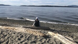 A person sitting on a rocky beach with a view of calm water and distant hills.