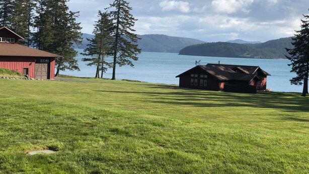 A red barn and a cabin by a lake with mountains in the background.