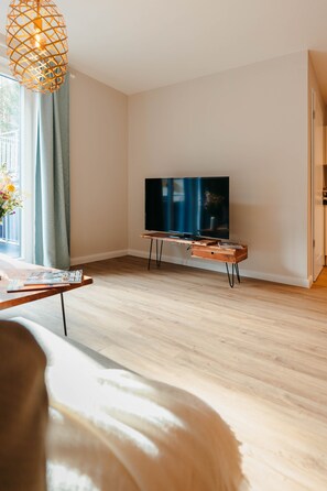 A modern living room with a wooden floor, a flat-screen TV on a wooden stand, a window with curtains, and a hanging light fixture.