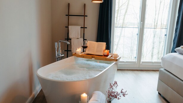 A modern bathroom with a freestanding bathtub, a wooden stool with a towel, and a window with curtains. - Apartment between forest and lake with free-standing bathtub and balcony (5 star DTV) (Ratzeburg)
