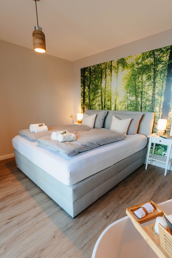 A modern bedroom with a large bed, a nightstand, a wall-mounted lamp, and a wooden floor.