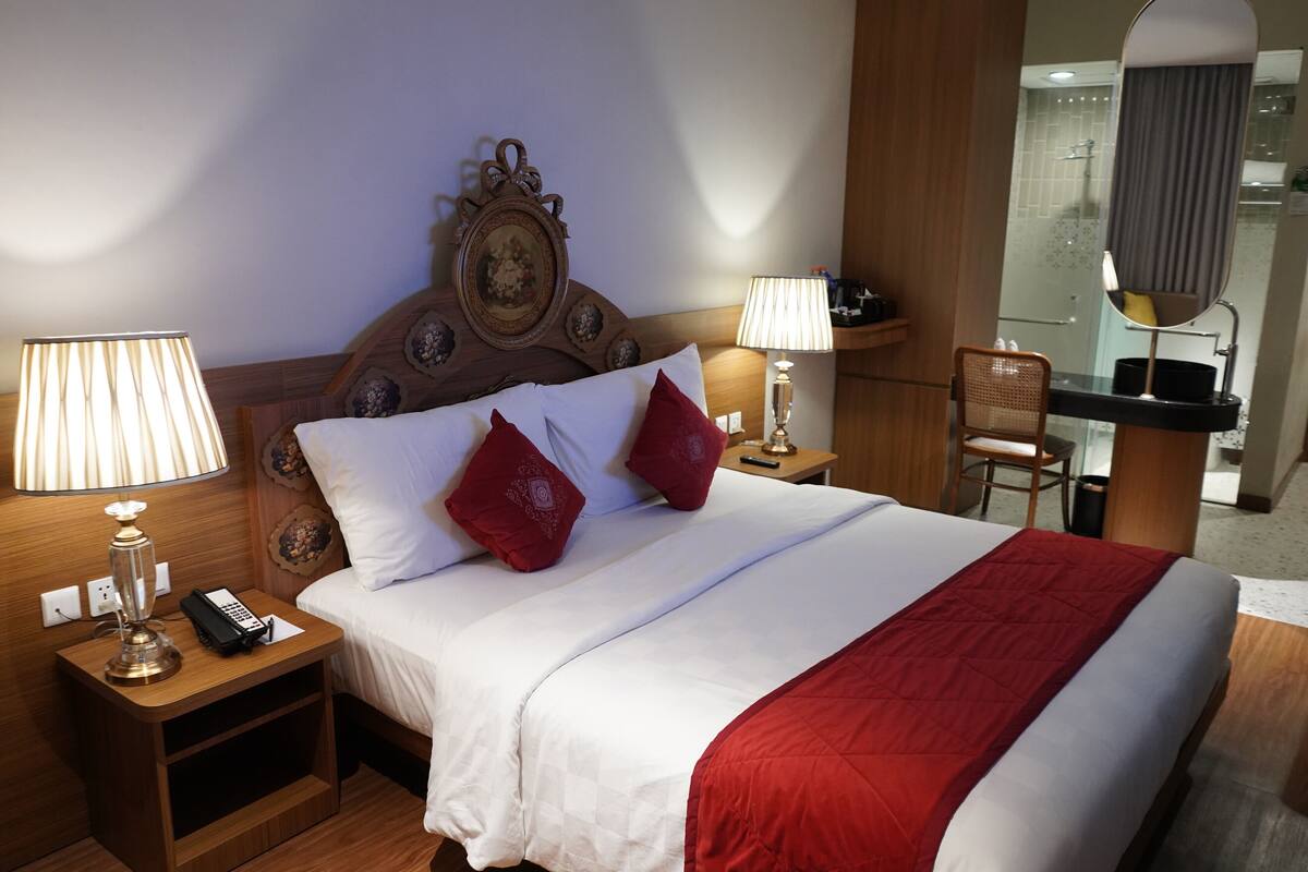 Quarto de hotel com cama, duas mesas de cabeceira, um criado-mudo, uma cadeira e banheiro visível através de um separador de vidro, além de um espelho.