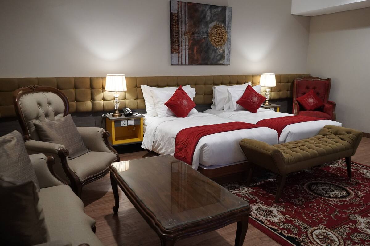 Quarto de hotel com uma cama grande, duas mesas de cabeceira, uma mesa de centro de madeira, um tapete com padrão dourado e vermelho, e uma poltrona vermelha.