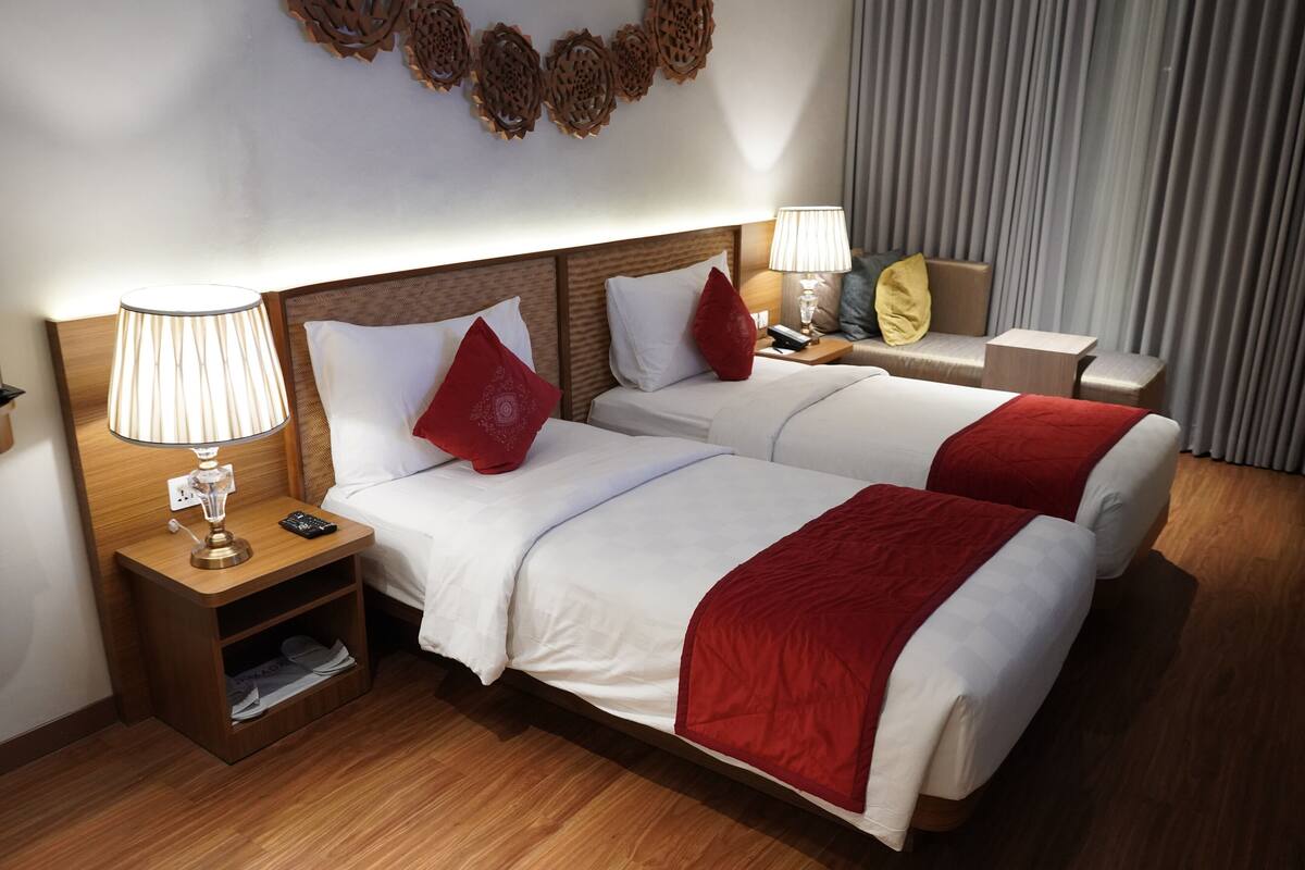 Quarto de hotel com duas camas, uma mesa de cabeceira com abajur, uma pequena mesa também com abajur e um objeto decorativo na parede.