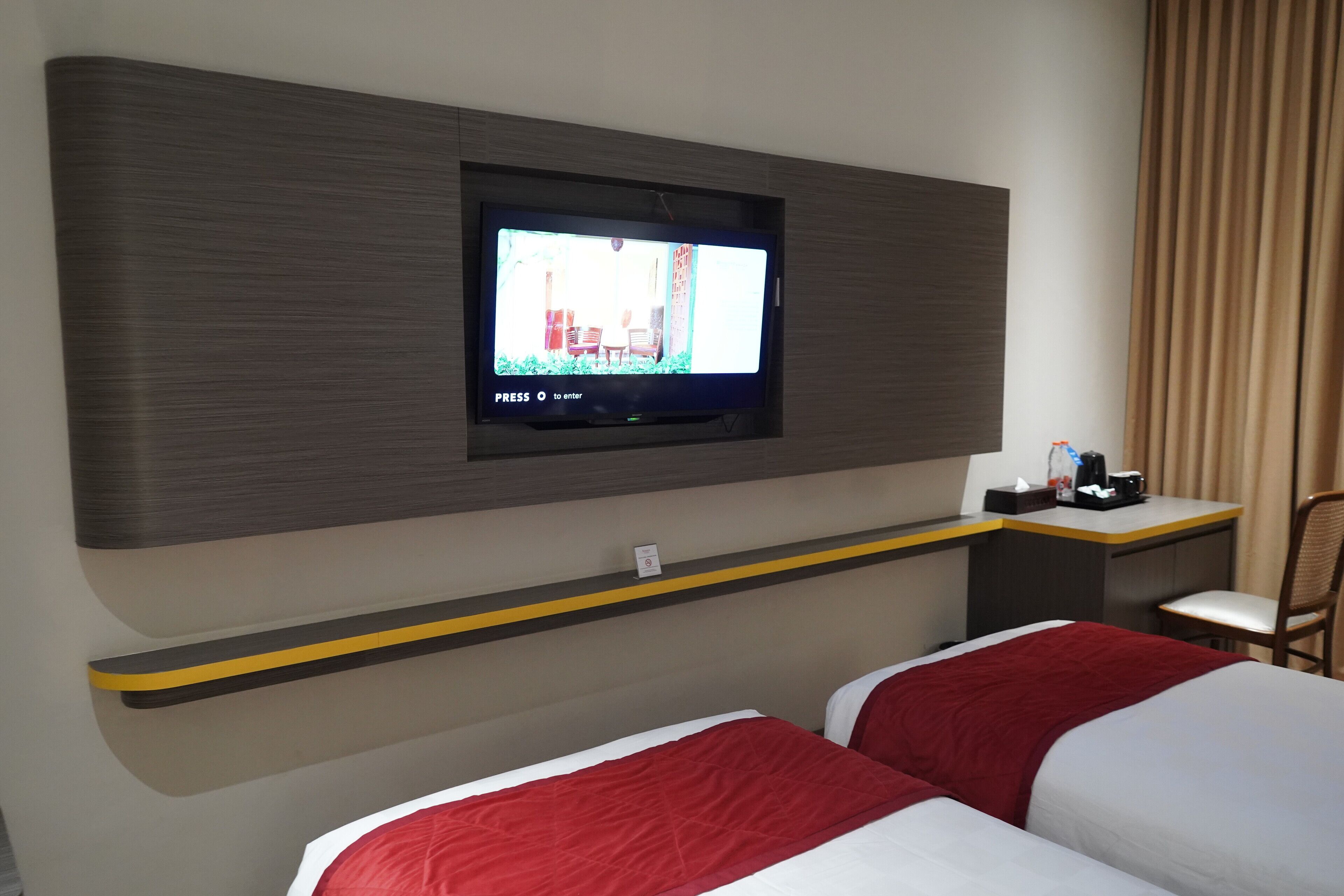 Ein Hotelzimmer mit an der Wand montiertem Fernseher, einem Schreibtisch mit Stuhl und zwei Betten mit rot-weißer Bettwäsche.