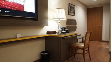 A hotel reception area with a TV screen displaying a message, a desk with a lamp, a trash bin, and a wooden chair.