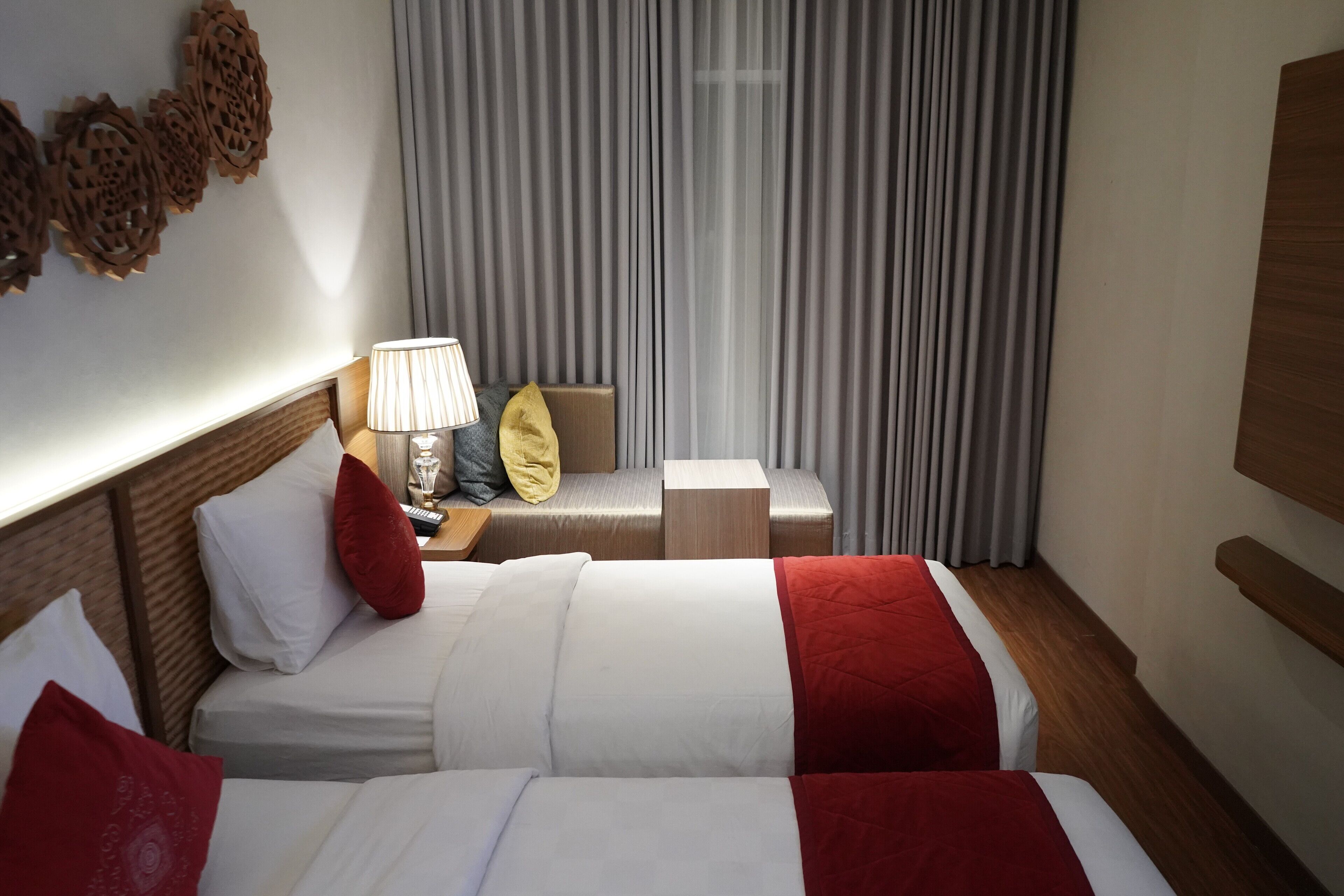 Habitación de hotel con dos camas, una mesita de noche con lámpara, cabecero de madera y una ventana grande con cortinas.