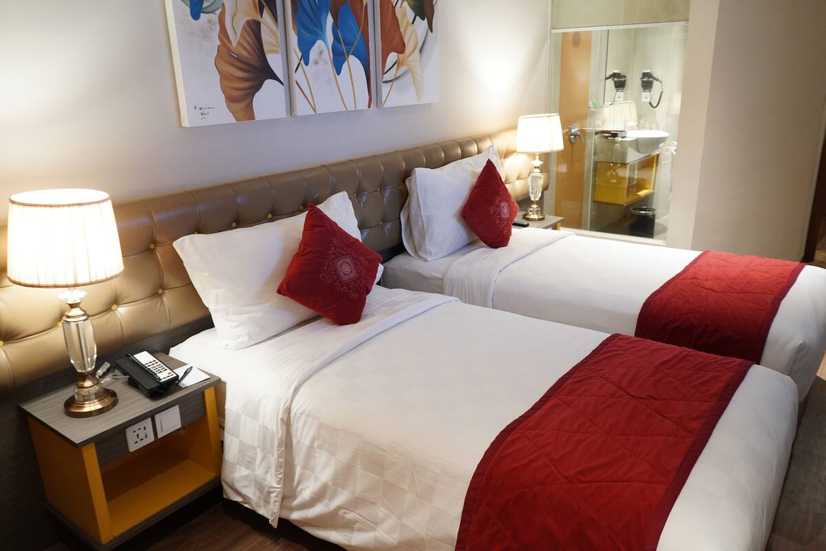 Quarto de hotel com duas camas, cada uma com roupa de cama branca e detalhes vermelhos, uma mesa de cabeceira com abajur e telefone, e uma obra de arte na parede.