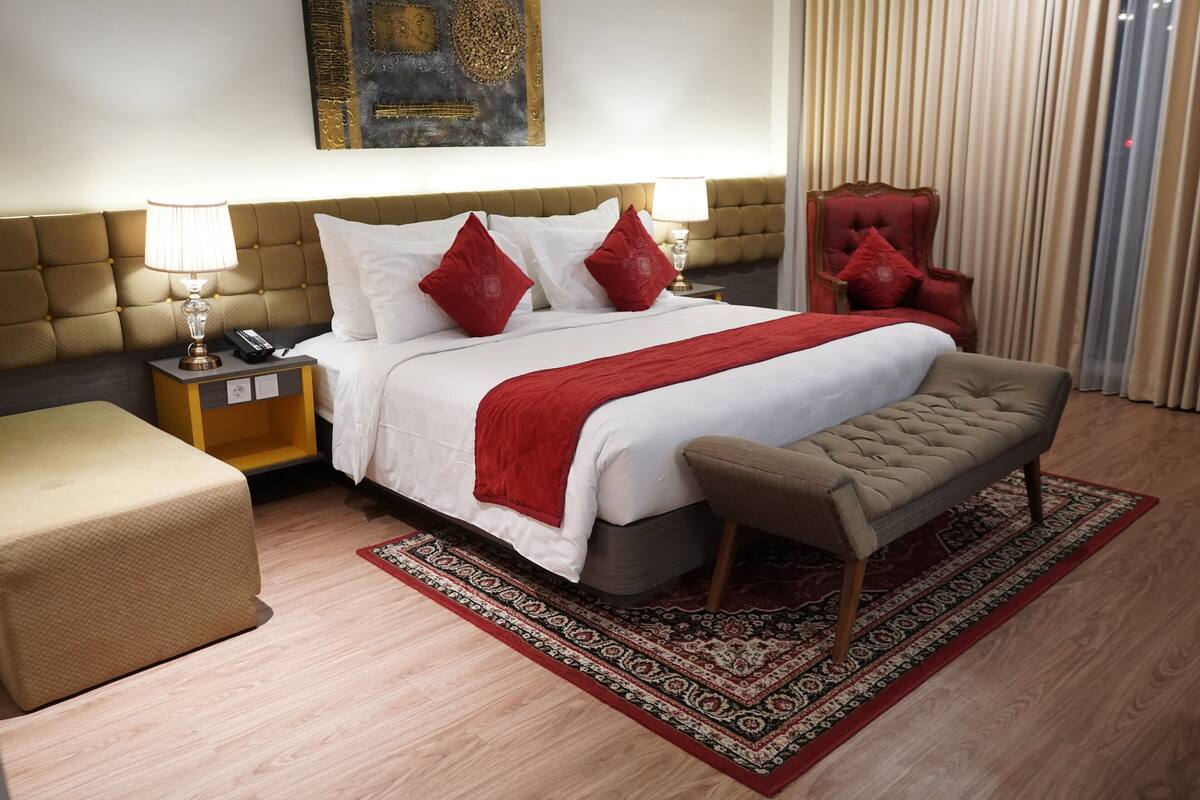 Quarto de hotel com uma cama grande, um banco, uma mesa de cabeceira, um abajur e uma cadeira.