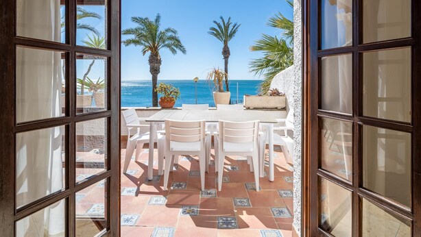 Une porte ouverte donne sur une terrasse offrant une vue sur la mer, avec une table, des chaises, des palmiers et des plantes en pot.