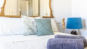Un letto rifatto con biancheria bianca, due cuscini fantasia e una lampada blu su un comodino.