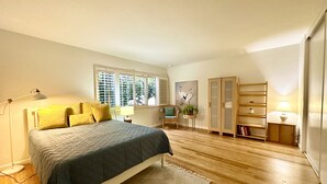 Ein Schlafzimmer mit einem Bett, einem Fenster mit Jalousien, einem Sessel, einem kleinen Tisch mit Lampe und einem Kleiderschrank.