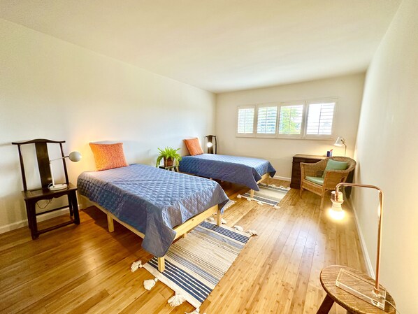 Habitación con dos camas, una silla, una mesita y una ventana con persianas.