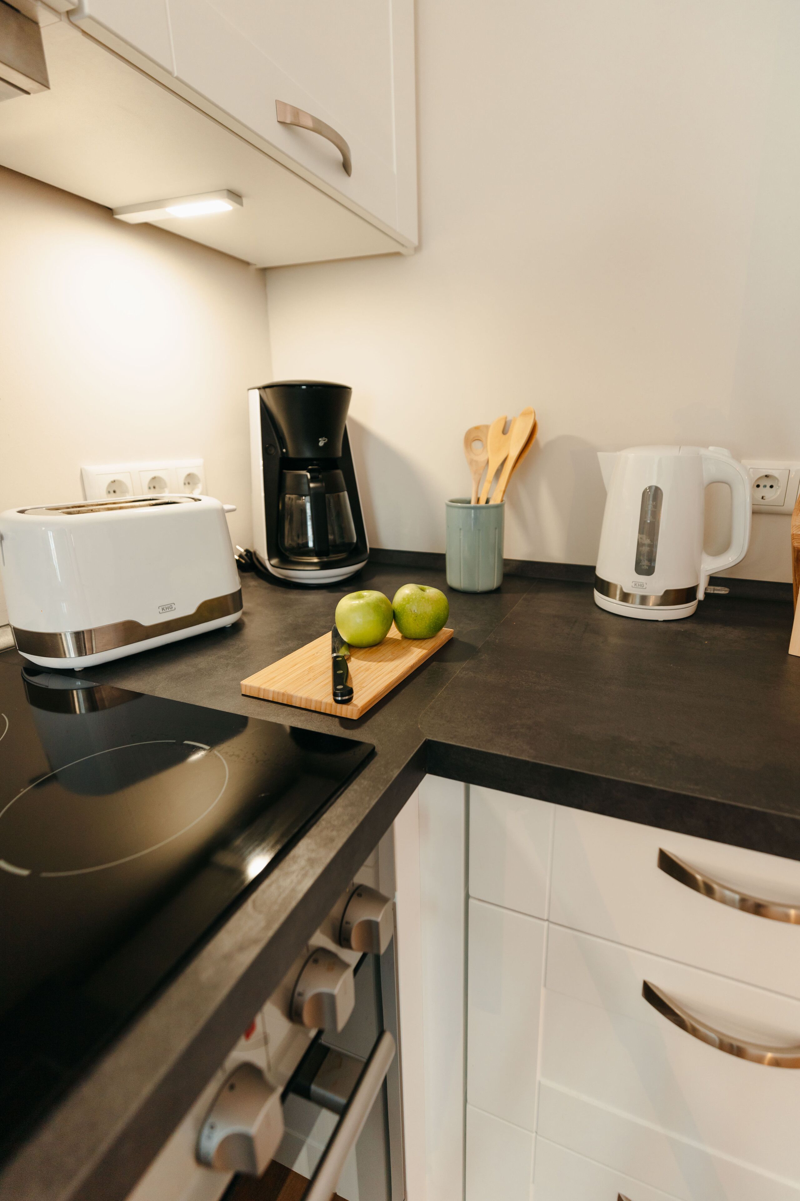 A kitchen with a toaster, coffee maker, kettle, and wooden cutting board with apples.