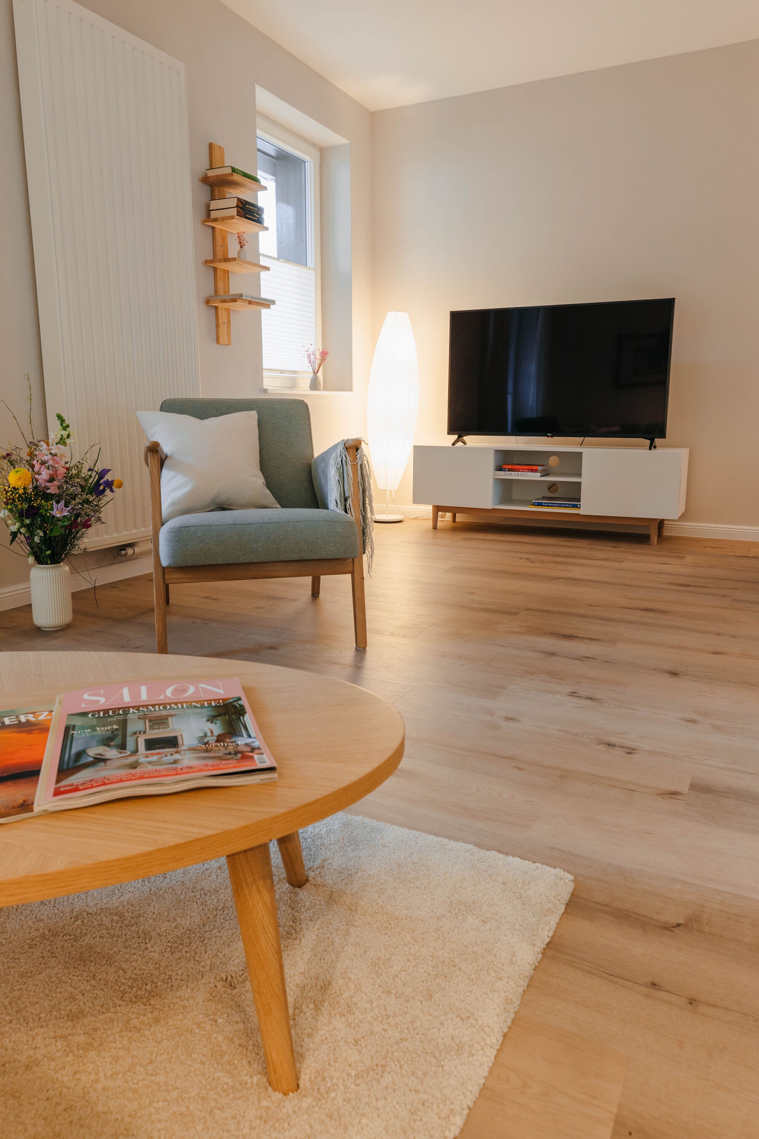 Un salon avec un plancher en bois, un fauteuil vert, un meuble TV blanc et un magazine sur une table basse.
