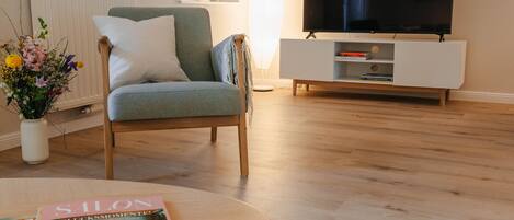 Un salon avec un plancher en bois, un fauteuil vert, un meuble TV blanc et un magazine sur une table basse.