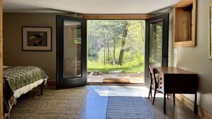 Ein Schlafzimmer mit Bett, Schreibtisch und Blick auf einen Wald durch geöffnete Türen.