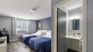 Une chambre d’hôtel avec deux lits, un bureau, un four à micro-ondes, une fenêtre avec des rideaux, une salle de bain avec un lavabo et un miroir, et une porte donnant sur une autre chambre.