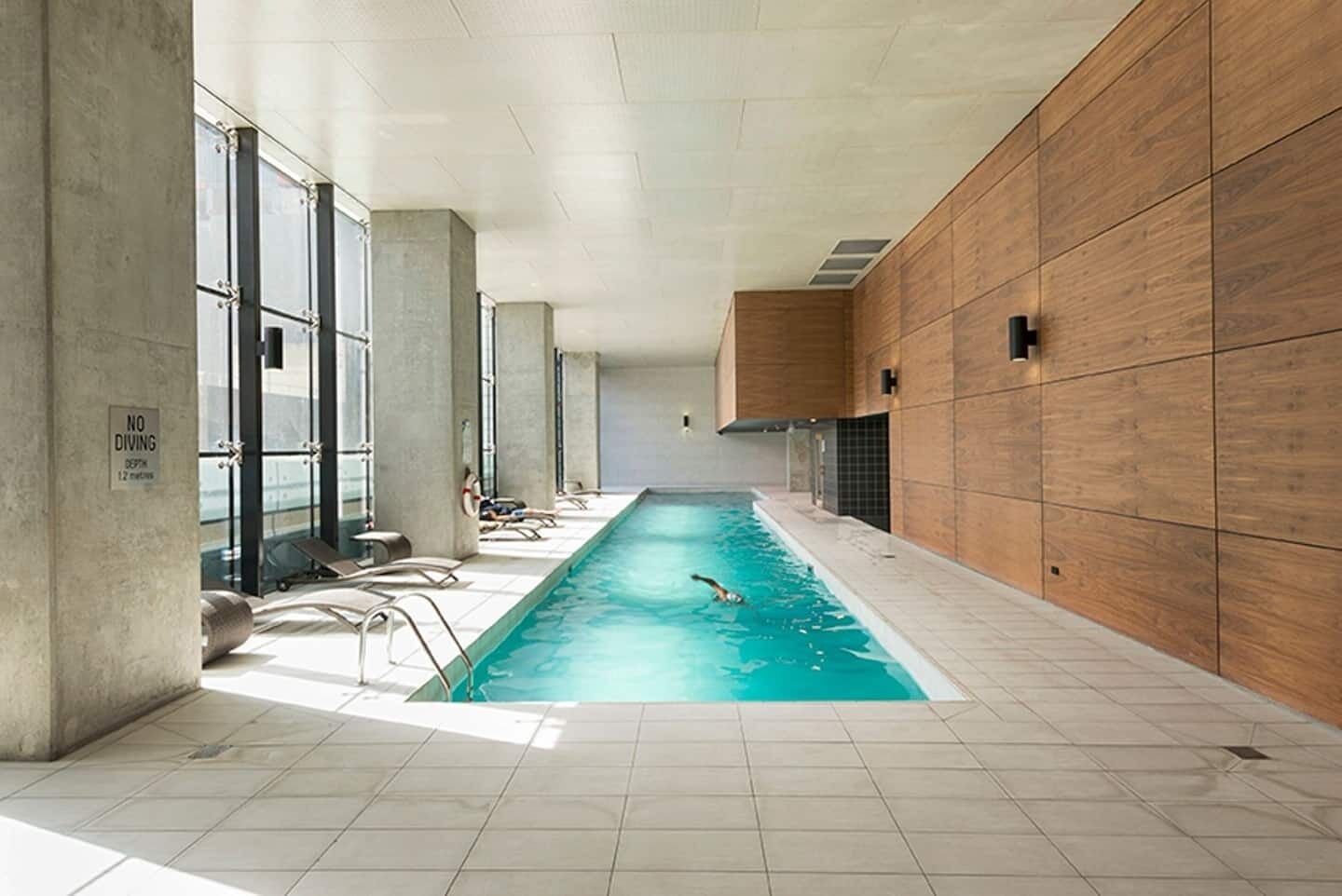 An indoor swimming pool with clear glass walls, a tiled floor, and a wooden wall on one side.