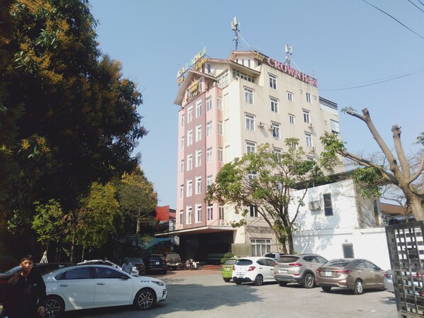 A multi-story hotel with a prominent sign, parked cars, and trees in the foreground. - Khach san Crown (Phan Dinh Phung)