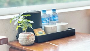 A tray with a rolled-up towel, a small potted plant, a bottle of water, a mug, and a packet of snacks on a wooden table by a window.