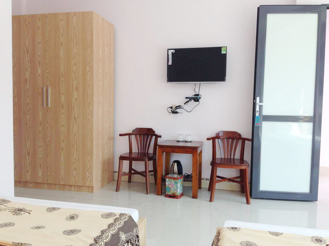 A room with a wooden wardrobe, a television mounted on the wall, two wooden chairs around a small table, and a cabinet with a glass door.